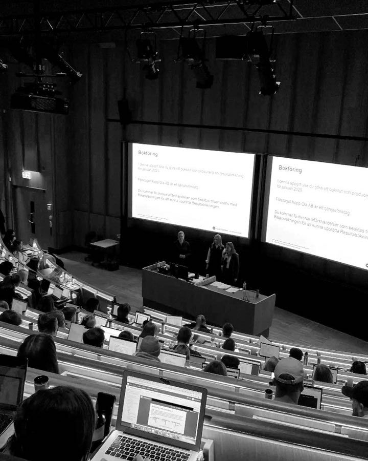 A lecture hall seen from the back row, two screens lit at the front.